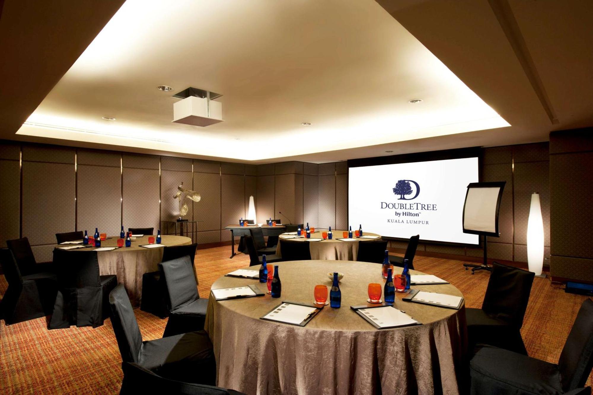 Doubletree By Hilton Kuala Lumpur Exterior foto The photo shows a conference room set up for a meeting or event. There are several round tables with beige tablecloths, each adorned with blue and red decorations, likely drinks and supplies. Black chairs are placed around the tables. On one wall, th